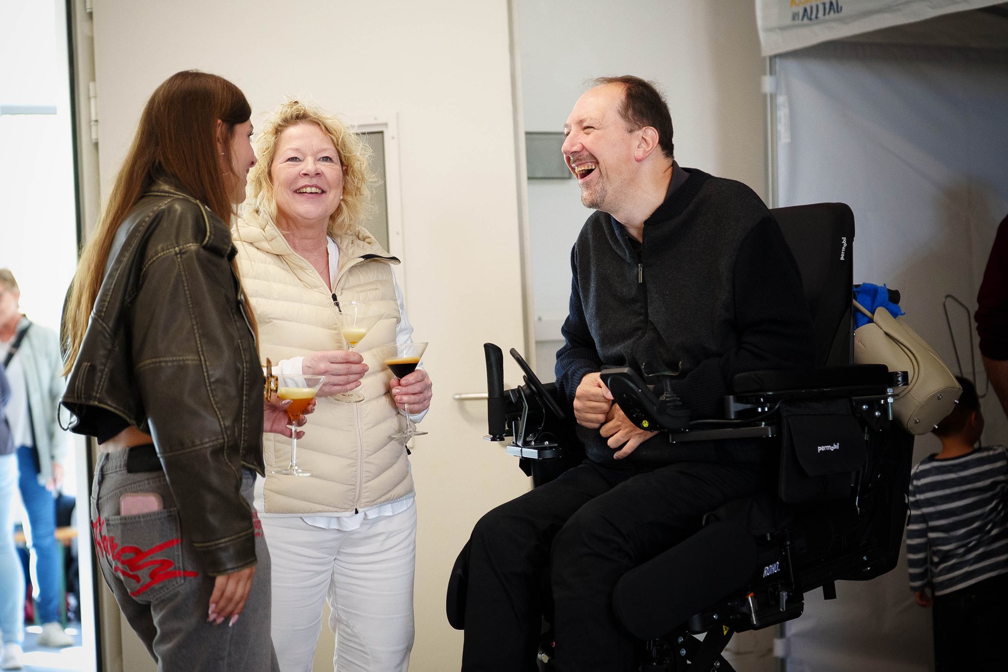 Gäste und ein Mann im Elektrorollstuhl lachen gemeinsam beim Sommerfest von Assistenten im Alltag